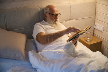 Photo for Old man with glasses reads a book before going to bed, he lies in a comfortable bed - Royalty Free Image