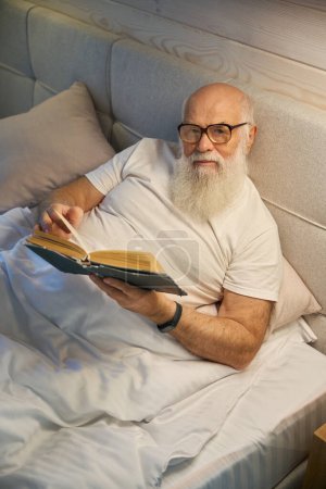 Téléchargez les photos : Vieil homme à barbe grise avec des lunettes lit un livre avant d'aller au lit, il se trouve dans un lit confortable - en image libre de droit