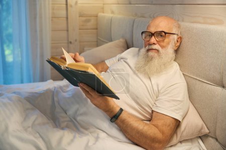 Téléchargez les photos : Vieil homme à barbe grise avec des lunettes lisant un livre avant d'aller au lit, il se trouve dans un lit confortable à la maison - en image libre de droit