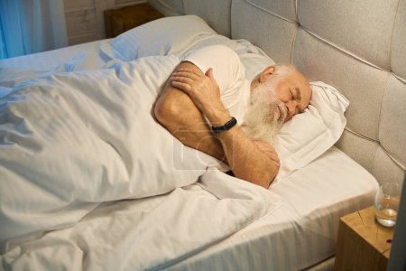 Téléchargez les photos : Homme âgé dort sur le côté dans un lit confortable, avec un verre d'eau sur la table de nuit à côté de lui - en image libre de droit