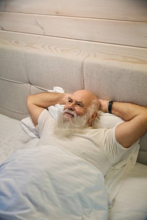 Téléchargez les photos : Vieil homme repose sur un lit confortable, la chambre a un intérieur minimaliste - en image libre de droit