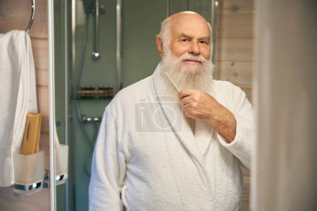 Téléchargez les photos : Le vieil homme aux cheveux gris peigne sa barbe devant un miroir, il est en peignoir blanc - en image libre de droit