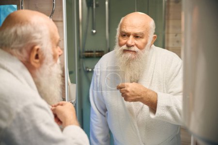 Foto de Viejo canoso haciendo su rutina matutina en el baño, está en un albornoz blanco - Imagen libre de derechos
