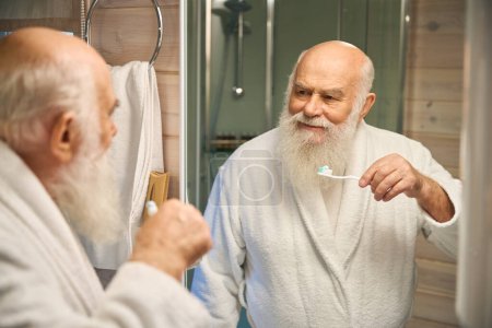 Téléchargez les photos : Vieil homme faisant sa routine matinale dans la salle de bain, tenant une brosse à dents - en image libre de droit