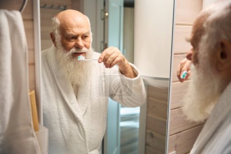 Téléchargez les photos : Le vieil homme se brosse les dents dans la salle de bain devant le miroir, il est dans un peignoir chaud - en image libre de droit