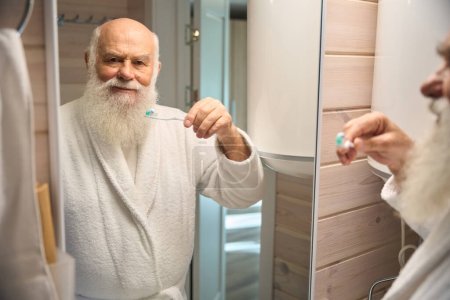 Téléchargez les photos : Heureux vieil homme se brosse les dents dans la salle de bain en face du miroir, il est dans un peignoir chaud - en image libre de droit