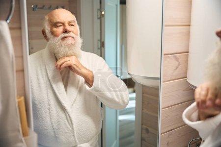 Téléchargez les photos : Vieil homme à barbe grise dans la salle de bain devant le miroir, il est dans un peignoir chaud - en image libre de droit