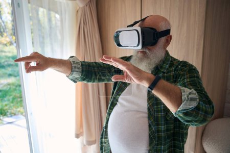 Téléchargez les photos : Homme âgé joue dans des lunettes de réalité virtuelle, il est dans une salle lumineuse confortable - en image libre de droit