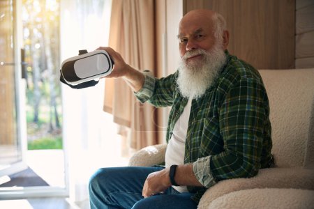 Téléchargez les photos : Heureux vieillard à barbe grise tient des lunettes de réalité virtuelle dans sa main, il est assis dans une salle lumineuse confortable - en image libre de droit