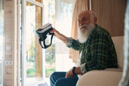 Foto de Viejo con una camisa a cuadros lleva gafas de realidad virtual en la mano, se sienta en una acogedora habitación luminosa - Imagen libre de derechos
