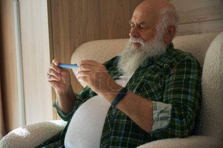 Photo for Old man in a checkered shirt holds a thermometer in his hand, he sits in a soft chair - Royalty Free Image