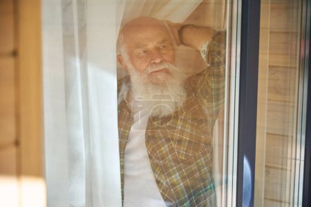 Téléchargez les photos : Le vieil homme en chemise à carreaux regarde par la fenêtre, il vit dans une maison en bois moderne - en image libre de droit