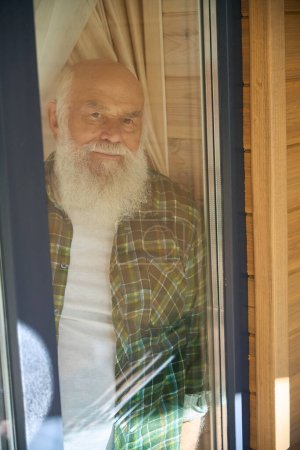 Téléchargez les photos : Un vieil homme aux cheveux gris souriant en chemise à carreaux regarde par la fenêtre, il vit dans une maison moderne en bois - en image libre de droit