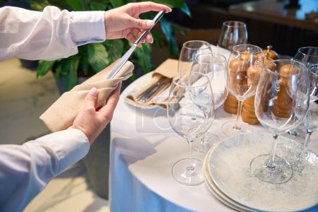 Téléchargez les photos : Serveur tient une serviette et des couverts dans sa main, il met la table dans la salle de restaurant - en image libre de droit
