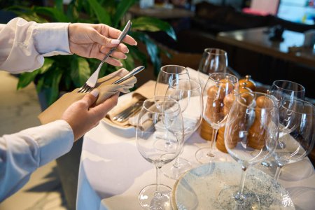 Photo for Waiter in white jacket holds a napkin and cutlery in his hand, he serves a table in a restaurant hall - Royalty Free Image