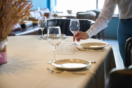 Foto de Camarera coloca un vaso grande junto al plato, hay un mantel blanco sobre la mesa - Imagen libre de derechos