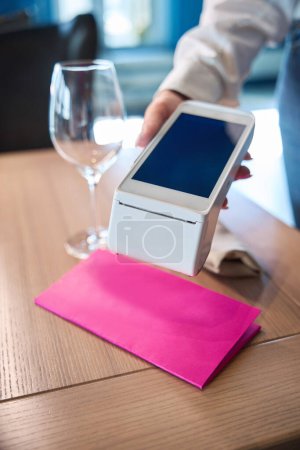 Téléchargez les photos : Femme tient un terminal pour payer les clients, sur la table il y a des couverts et une enveloppe rose avec une facture - en image libre de droit