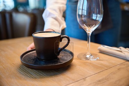 Foto de Camarera pone una taza de café en una mesa de madera, hay un vaso grande en la mesa - Imagen libre de derechos