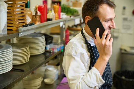 Photo for Cook in a chefs apron communicates on his mobile phone, there is creative chaos in the workplace - Royalty Free Image