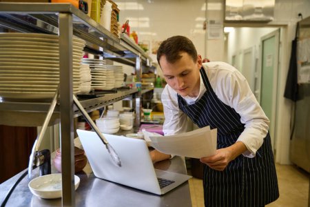 Photo for Cook is working with documents at the kitchen rack, he is using a laptop - Royalty Free Image