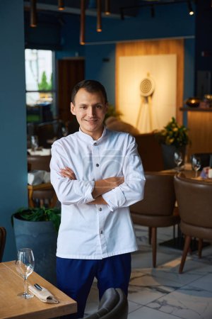 Téléchargez les photos : Chef souriant dans un uniforme de chefs se tient dans une salle de restaurant, intérieur intérieur moderne - en image libre de droit