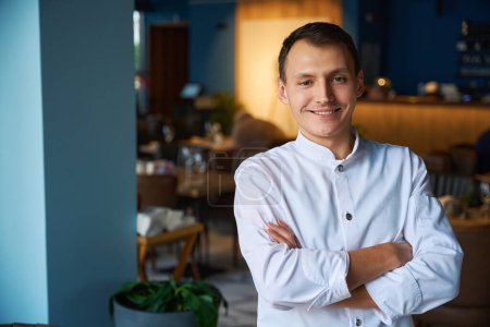 Photo for Young chef in a chefs uniform stands in a restaurant hall, indoors modern interior - Royalty Free Image