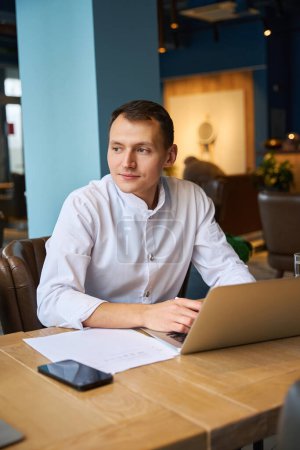 Téléchargez les photos : Homme en uniforme de cuisine travaille avec des documents dans la salle de restaurant, il utilise un ordinateur portable - en image libre de droit
