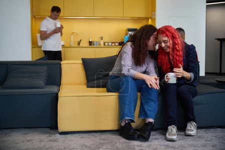 Téléchargez les photos : Deux filles chuchotant au repos et buvant du café sur un canapé doux pendant la pause au bureau, bavardant sur leurs collègues et leur vie privée, espace de coworking - en image libre de droit