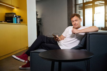 Téléchargez les photos : Beau jeune homme buvant du café et surfant sur internet sur son smartphone assis sur un canapé douillet, travail indépendant dans un espace de coworking, technologies modernes - en image libre de droit