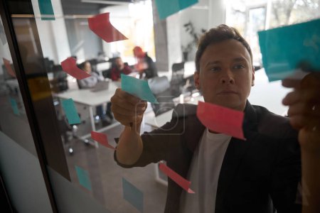 Photo for Concentrated business man looking through memo notes stickered to the transparent wall of office room, team of managers keeping brainstorm, suggesting ideas - Royalty Free Image