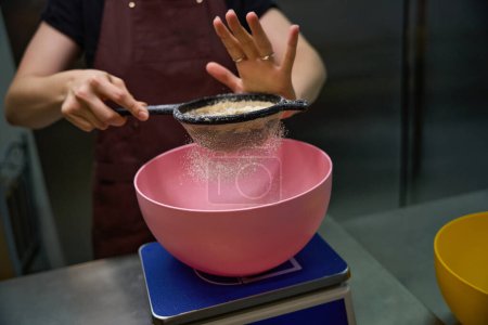 Foto de Foto recortada de pastelero en delantal tamizar harina de almendras a través de tamiz sobre tazón poner en escala de cocina - Imagen libre de derechos