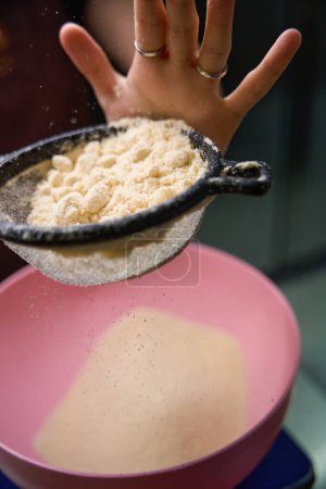 Téléchargez les photos : Gros plan de la main féminine tamiser la farine d'amande à travers un tamis sur un bol placé sur une balance de cuisine - en image libre de droit