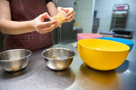 Foto de Foto recortada del chef pastelero en delantal separando la clara de huevo de la yema sobre el tazón en la mesa - Imagen libre de derechos
