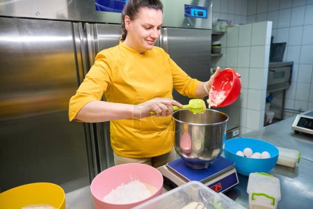 Téléchargez les photos : Boulanger féminin souriant ajoutant de la pâte au bol placé sur la balance sur la table - en image libre de droit