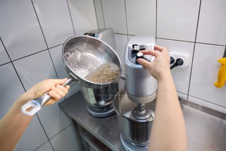 Photo for Closeup of female pastry chef hands pouring sugar syrup from saucepan into mixing bowl - Royalty Free Image
