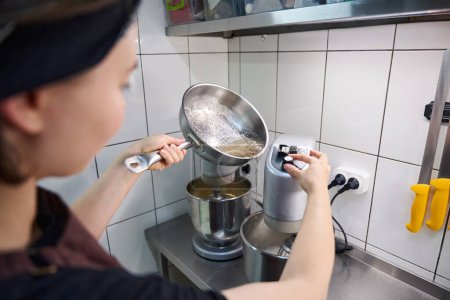 Photo for Female confectioner pouring sugar syrup from saucepan into mixing bowl on kitchen countertop - Royalty Free Image