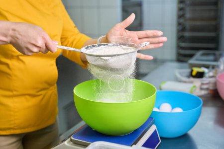 Foto de Foto recortada de cocinero en uniforme tamizar la harina a través de tamiz sobre un tazón colocado en la escala de la cocina en la mesa - Imagen libre de derechos