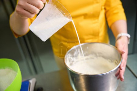 Téléchargez les photos : Photo recadrée d'un boulanger versant du lait de la tasse à mesurer dans un bol avec de la pâte - en image libre de droit