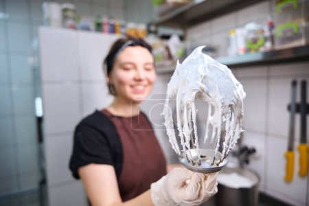 Téléchargez les photos : Agréable jeune boulanger femelle tenant fouet avec des blancs d'œufs fouettés devant la caméra - en image libre de droit