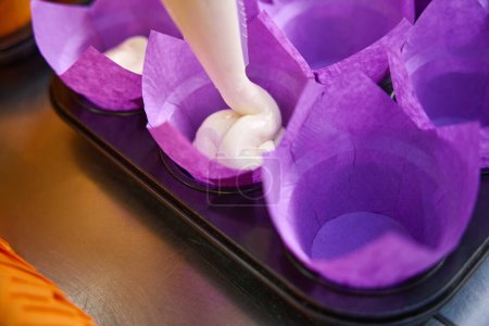 Téléchargez les photos : Gros plan de pâte serrée à travers la pointe du sac de tuyauterie en plastique dans des doublures de cupcake violet sur un plateau de muffins en métal sur le comptoir de la cuisine - en image libre de droit