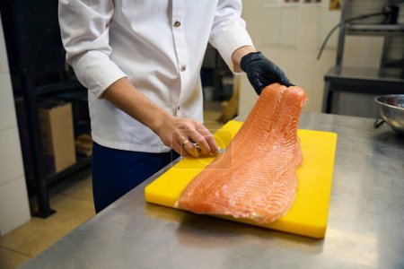 Téléchargez les photos : Cook coupe du poisson rouge avec un couteau tranchant, il travaille sur une grande planche à découper jaune - en image libre de droit