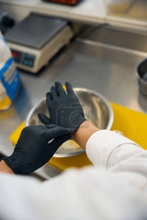 Téléchargez les photos : Travailleur de cuisine met des gants de protection noirs, il y a un bol et une planche à découper sur la table de travail - en image libre de droit
