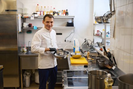 Photo for Smiling young chef preparing food in a restaurant kitchen, he is in a comfortable uniform - Royalty Free Image
