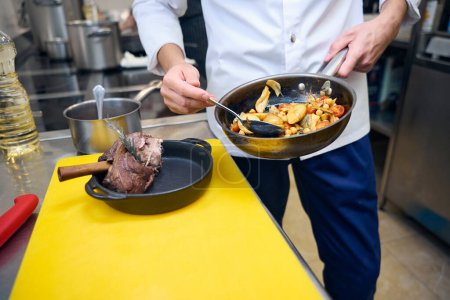Téléchargez les photos : Employé de cuisine prépare un jarret d'agneau avec des légumes et de la sauce pour servir, il utilise des produits de haute qualité - en image libre de droit