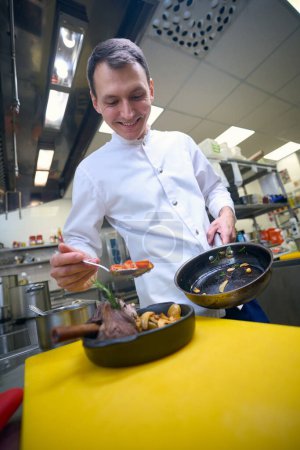 Téléchargez les photos : Jeune chef se prépare à servir jarret d'agneau avec des légumes, il prépare la nourriture dans la cuisine du restaurant - en image libre de droit