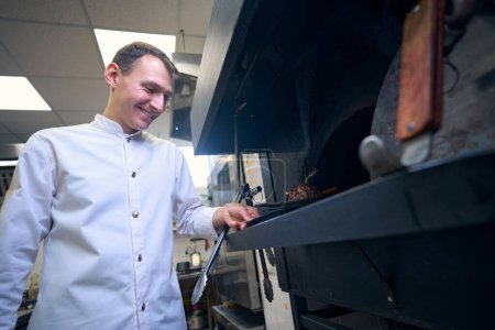 Foto de Chef sonriente pone una sartén con carne en el horno, utiliza productos de calidad - Imagen libre de derechos