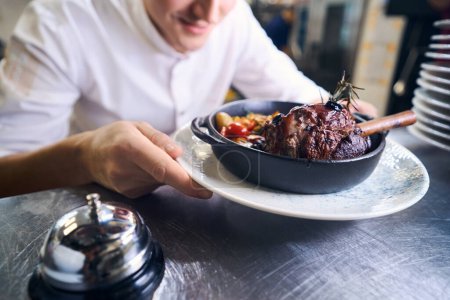 Photo for Young kitchen employee has prepared lamb shank with vegetables for serving, he is preparing food in the restaurant kitchen - Royalty Free Image