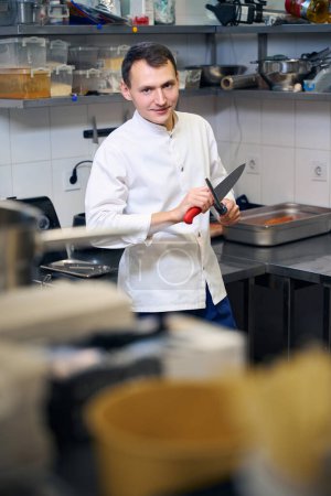 Téléchargez les photos : Travailleur de cuisine aiguise un grand couteau professionnel, il ya beaucoup d'équipement de cuisine autour - en image libre de droit