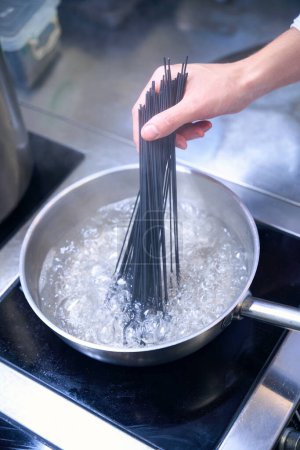 Foto de El hombre pone una porción de espagueti negro en agua hirviendo, usa una estufa moderna - Imagen libre de derechos