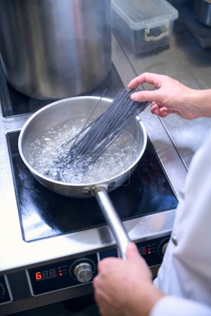 Cook puts a portion of black spaghetti into boiling water, he uses a modern stove